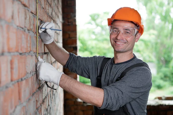 close-up of a caucasian builder in a protective helmet and glasses measures a brick wall with a tape measure at a construction site. Measurements of the height of the wall. Construction concept.