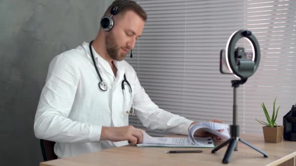 En läkare i vit rock sitter vid ett bord på ett kontor och undersöker en avlägsen patient. Läkaren talar med klienten med hjälp av en virtuell chattprogram. Begreppet distansvård. — Stockvideo