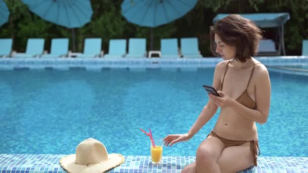 In summer, while on vacation at a resort at the edge of the pool, a Caucasian woman in a swimsuit sits reading a news feed on her smartphone and drinks freshly squeezed juice. — Stock Video