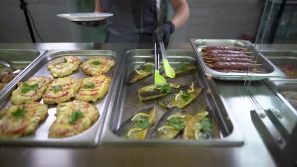 En una cafetería escolar o buffet en la línea de servicio de comida, los asistentes con guantes negros colocan pescado horneado con limón y caballa en un plato. — Vídeos de Stock