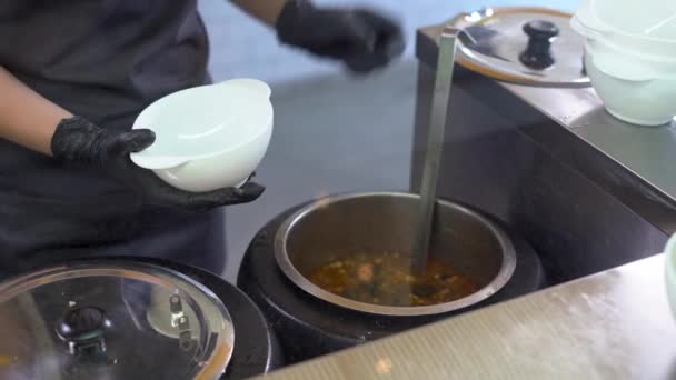 En una cafetería escolar o en un buffet de comida, el personal de servicio de guantes negros sirve un plato de caldo o sopa caliente y fresca.. — Vídeos de Stock