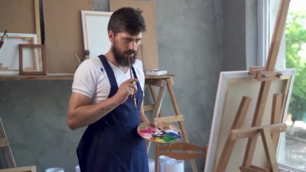 A young bearded Caucasian male painter dressed in a blue apron stands near an easel with a canvas holding a palette with paints in his hands and thinks about what to draw. — Wideo stockowe