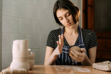 Uzun siyah saçlı, genç ve karizmatik bir kadın evde oturup kilden bir çömlek yontuyor. Ellerinde büyük bir parça kil yoğuruyor..
