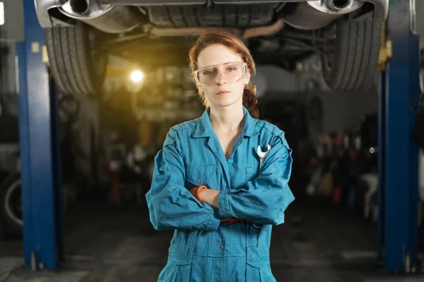 A young woman in work clothes, an apprentice stands next to the car and is proud and happy in the garage. holding a wrench in his hands. Copy space.
