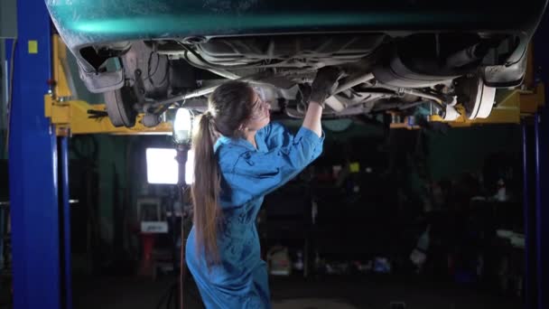 Kaukasische junge Automechanikerin in Overalls, die unter einem Fahrzeug in einem Autoservice arbeitet. Die Frau trug Handschuhe unter dem Auto. Authentische echte Werkstatt. — Stockvideo