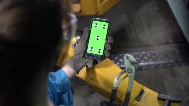 Vista del hombro de una mujer mecánico de coche en gafas y monos con un teléfono inteligente en la mano y mirando a una pantalla verde con una tecla de croma. Material de vídeo. Teléfono móvil. — Vídeos de Stock