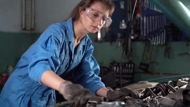 Mujer caucásica mecánico de automóviles se encuentra en overoles y gafas cerca de la capucha abierta del coche. reparación del motor. Desenrosque con llaves. — Vídeos de Stock