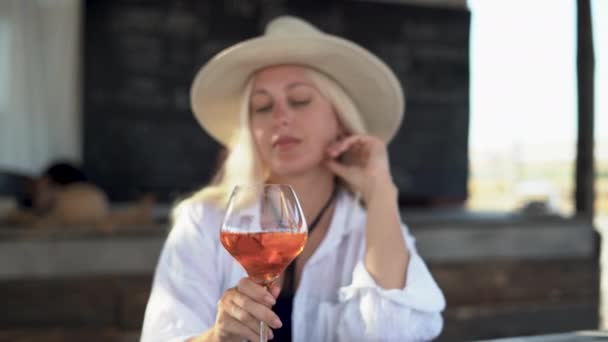 Young beautiful woman in casual clothes and a straw hat sits at a bar at the resort and drinks a refreshing cocktail. — Stock Video