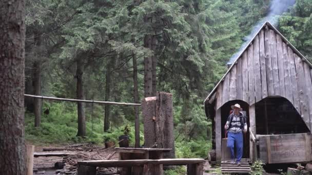 Stary dom leśnika w lesie, dym pochodzi z komina, biały mężczyzna turysta z laskami i plecakiem wychodzi z chaty i oddycha świeżym powietrzem. — Wideo stockowe