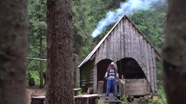 Ormandaki ormancının eski evi, bacadan duman çıkıyor, verandada duran beyaz erkek turist sırt çantasını çıkarmaya başlıyor. Tatil kavramı — Stok video