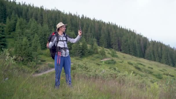 Verklig vit man turist med en ryggsäck och pinnar för promenader stiot på en kulle nära skogen och gör ett videosamtal, kommunicera med släktingar och vänner. — Stockvideo