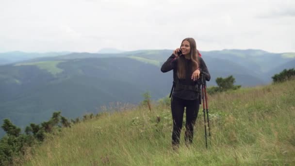 En ung kvinna med ryggsäck på axlarna står på toppen av ett berg på hösten eller våren, håller i en käpp och kommunicerar med någon på sin smartphone. vandringskoncept. — Stockvideo