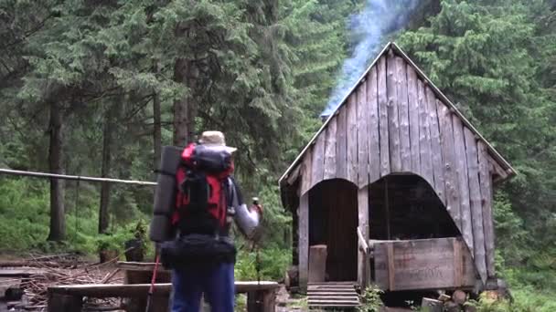 La antigua casa del guardabosques en el bosque, el humo proviene de la chimenea, un turista masculino caucásico con bastones y una mochila camina a través del bosque hacia la cabaña. Concepto de vacaciones — Vídeos de Stock