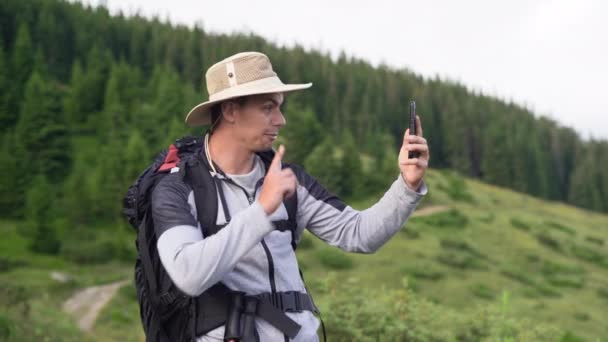 Close-up portret van een echte blanke mannelijke toerist met een rugzak en stokken voor het lopen stiot op een heuvel in de buurt van het bos en maakt een video gesprek, communiceren met familieleden en vrienden. — Stockvideo