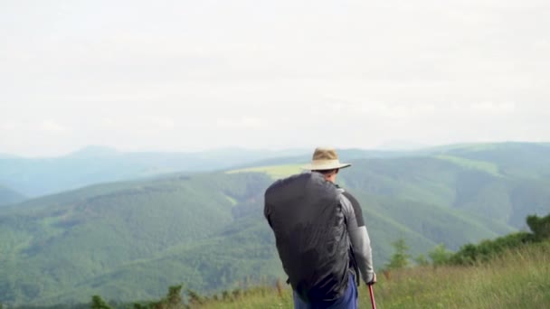 Vista trasera de un turista con una gran mochila en las montañas mira a los picos. hombre caminando bajo la lluvia con una cubierta protectora en su mochila. Concepto de viajes y turismo. — Vídeos de Stock