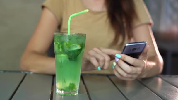 Ein Glas alkoholischen Mojito auf einem Tisch in einem Café Mädchen mit einem Telefon chatten online trinken einen exotischen Cocktail. Entspannung auf der Straße am Tisch. Konzept für Sommergetränke. — Stockvideo