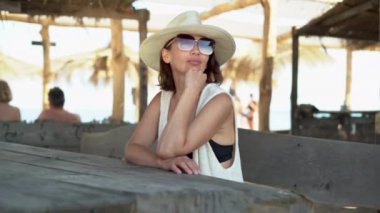 Şapkalı ve gözlüklü beyaz kadın tropikal bir barda oturmuş kokteyl bekliyor. Garson kız müşteriye içki getiriyor..