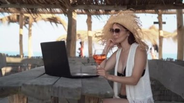 Hasır şapkalı güzel bir beyaz kadın tropik bir barda oturmuş kokteyl içiyor ve bilgisayarını ve uygulamasını kullanarak video görüşmesi yapıyor..