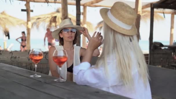 Dwie piękne kaukaskie kobiety w kapeluszach siedzą w kawiarni w kurorcie podczas wakacji na tropikalnej plaży i rozmawiają ze sobą. — Wideo stockowe