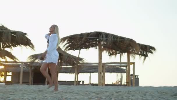 Een jonge mooie vrouw op vakantie wandelt langs een tropisch strand in de buurt van stroparasols houdt een koptelefoon in haar handen en geniet van het leven op een paradijselijk eiland in een resort. — Stockvideo