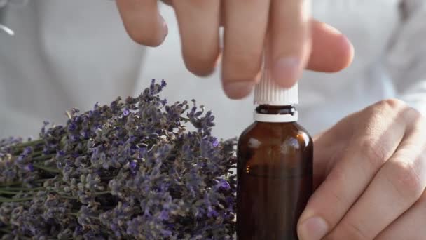 Una mano del hombre sostiene una pipeta de una botella con una planta pequeña. Flores de lavanda se encuentran cerca. Frascos cosméticos para aceite, aceites esenciales líquidos, suero de colágeno. — Vídeos de Stock