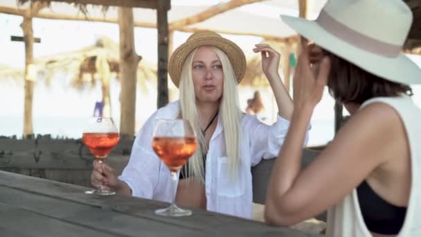 Dwie piękne kaukaskie kobiety w kapeluszach siedzą w kawiarni w kurorcie podczas wakacji na tropikalnej plaży i rozmawiają ze sobą. wznieśmy toast i uśmiechnijmy się radośnie. — Wideo stockowe
