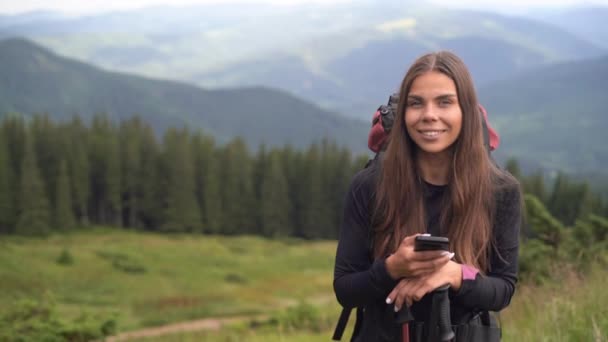 Une belle femme caucasienne est debout sur le sommet d'une montagne regardant la caméra et souriant. En arrière-plan, il y a des sommets de montagne, de beaux paysages. — Video