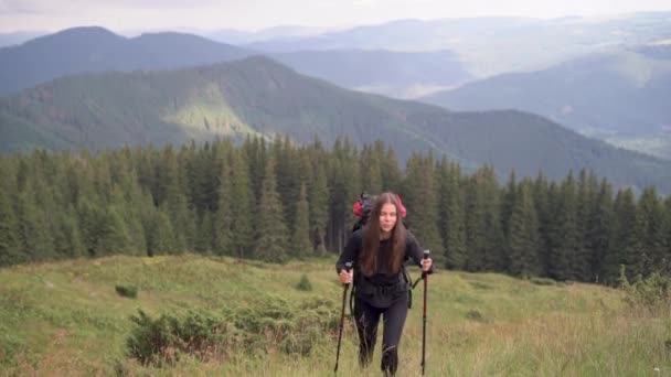 En ung kaukasisk kvinna på semester vandrar på en bergsrygg. Med pinnar klättrar en nordisk promenad upp på toppen av berget och ler. Begreppet aktiv vila. — Stockvideo