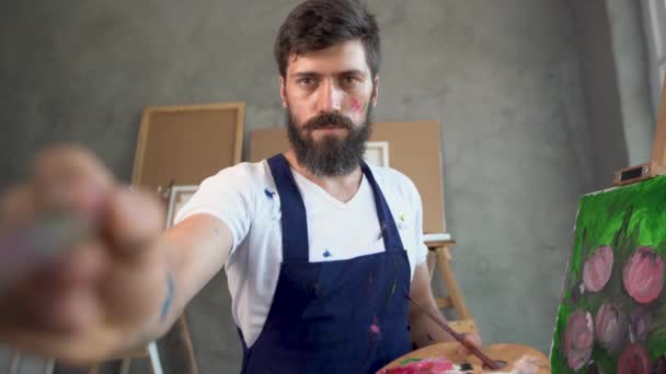 Dans un atelier d'art, un artiste masculin, vêtu d'une blouse de laboratoire avec une barbe, se tient debout et agite un pinceau. le concept brosse un tableau. — Video