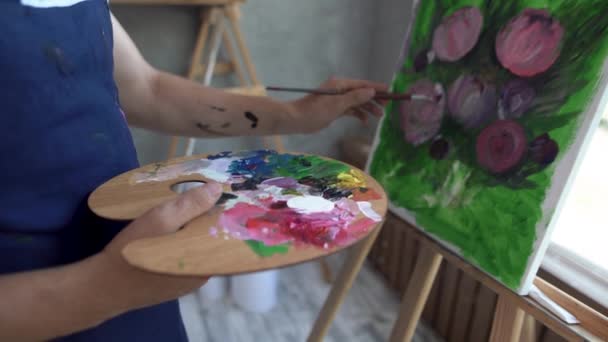 Primer plano de las manos masculinas en una paleta mezclar diferentes pinturas y flores de pintura sobre lienzo con un pincel. — Vídeo de stock