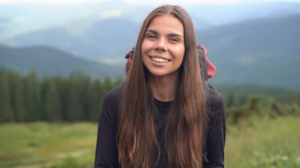 Genç beyaz bir kadının yakın plan portresi, dağ zirvelerinin arka planında duruyor, kameraya bakıyor ve gülümsüyor.. — Stok video