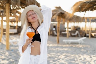 Beyaz gömlekli, tropik bir plajda elinde alkolsüz bir kokteyl olan bir bardağın altında duran sarışın güzel bir kadın gözlerini kapadı. Boşluğu kopyala.