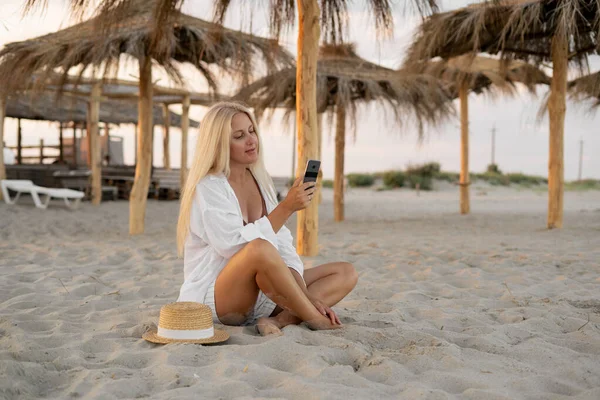 ビーチで休暇中に砂の上に座っているセクシーな白人女性は わら傘の下で彼女のスマートフォンを使って友人とビデオ通信を共有しています スペースのコピー — ストック写真