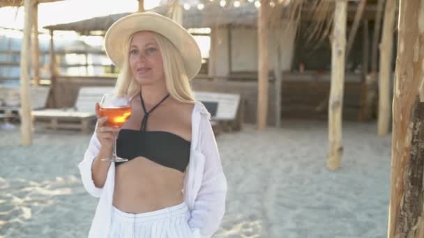 Krásná žena stojí na tropické pláži v blízkosti oceánu, drží v rukou koktejl a užívá si západu slunce. — Stock video