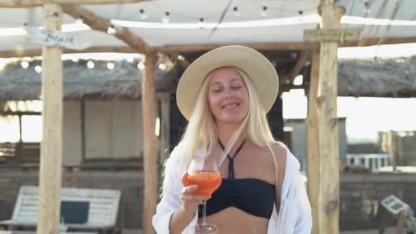 Een mooie blanke vrouw loopt tussen de stroparasols op het strand en houdt een alcoholische cocktail in haar hand. Vakantie concept op tropisch resort tijdens vakantie. — Stockvideo