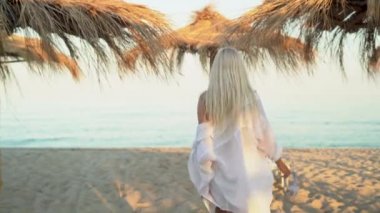 Güzel sarışın bir kadın kumlu sahil boyunca saman şemsiyeler arasında denize doğru yürüyor. Tropik bir tatil beldesinde tatil kavramı.