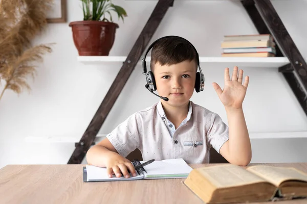 Pupil in wireless headphones with a microphone communicates on a webcam with a teacher watching a webinar or conference. The child studies at home remotely in the zoom. Online education concept.