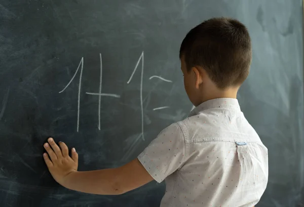 schoolboy in the class near the blackboard writes numbers with chalk. Math lesson. Junior School. The concept of modern education. Back to school. Place for text.