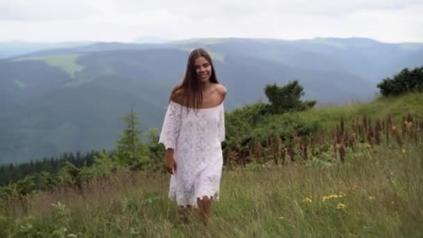 Una joven con un vestido blanco camina sobre un campo de hierba en una noche ventosa en las montañas de otoño con los pies desnudos. — Vídeos de Stock