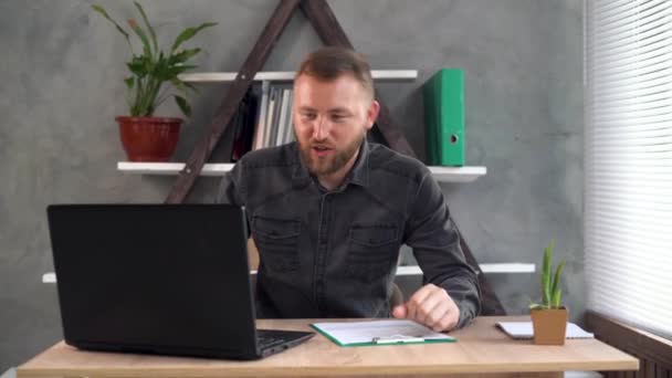 Um homem de negócios barbudo senta-se em sua mesa em casa em seu escritório, faz uma chamada de vídeo, mostra ao cliente um tablet com formulários preenchidos e diz algo. — Vídeo de Stock