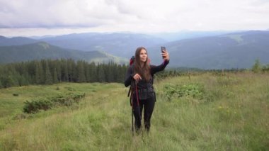 Dağlardaki tepede, büyük bir turist sırt çantası ve yürüyüş direkleri olan bir kadın var, elinde bir akıllı telefon tutuyor ve 6G internet ve bir uygulama kullanarak bir video görüşmesi yapıyor..
