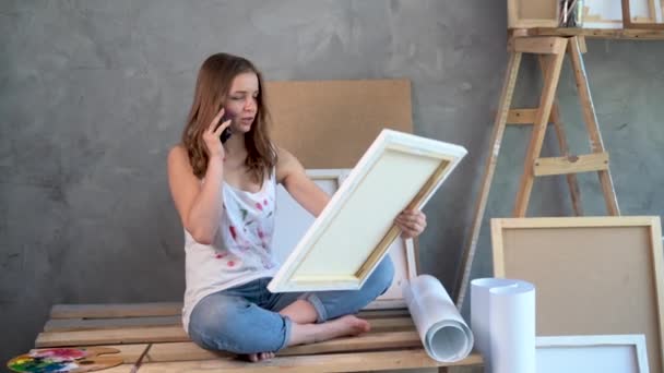 Une femme artiste caucasienne assise dans un studio d'art regardant une peinture et bavardant sur un téléphone portable. — Video
