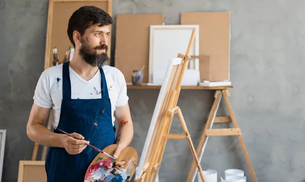 A bearded male artist dressed in a blue dirty apron stands in an art studio holding a palette with paints in his hands looks at the empty space for the text.