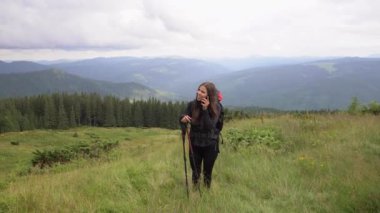 A beautiful Caucasian woman with a large backpack on her shoulders stands on a mountain ridge and has a conversation with someone on her smartphone.