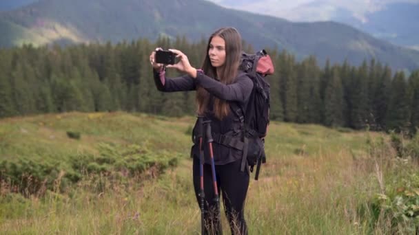 En ung vit kvinna vandrare medan vandring i bergen med en ryggsäck står på en bergsrygg håller sin smartphone i händerna och tar ett foto av landskapet. — Stockvideo