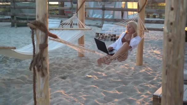 Молода жінка працює на ноутбуці, лежачи в гамаку на піщаному пляжі тропічного острова. Концепція зовнішньої позаштатної роботи . — стокове відео