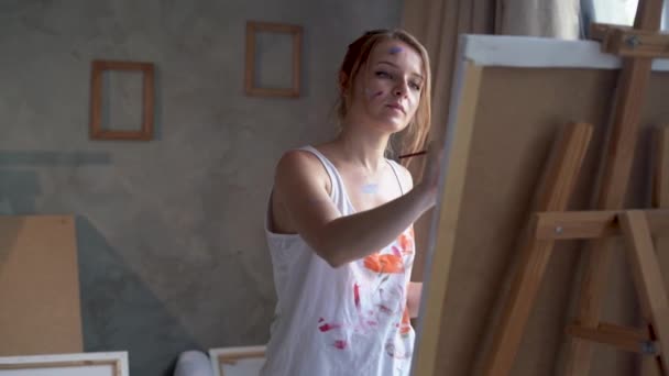 One woman artist stands near an easel in an art studio holding a brush in her hand and paints a picture. — Video