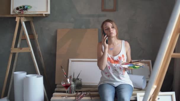 One young woman student artist holds a palette with paints in her hands and talks to someone through her smartphone sitting in an art studio. Copy space. — Vídeo de stock