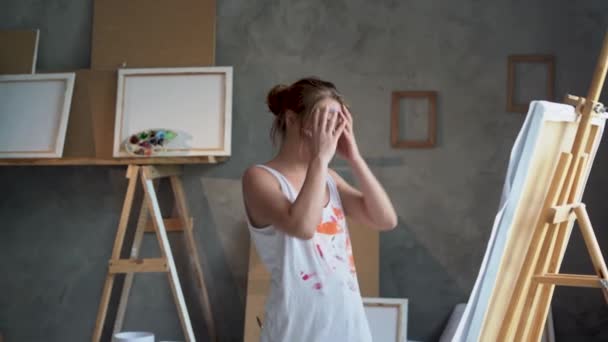 The depressed woman of the artist stands near the easel in the art studio, tears off a leaf on the started painting and throws it away. — Video