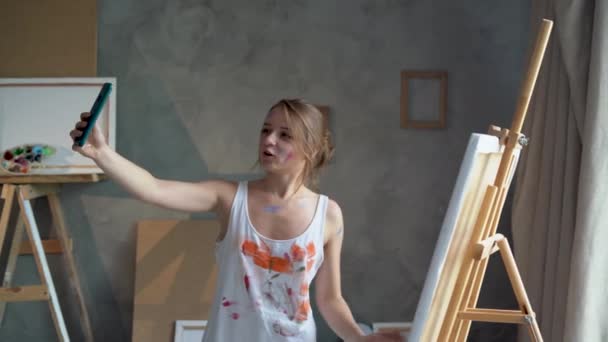 A young beautiful woman artist stands in an art studio communicates with someone on the phone video communication through the application, shows her picture. — Stock video